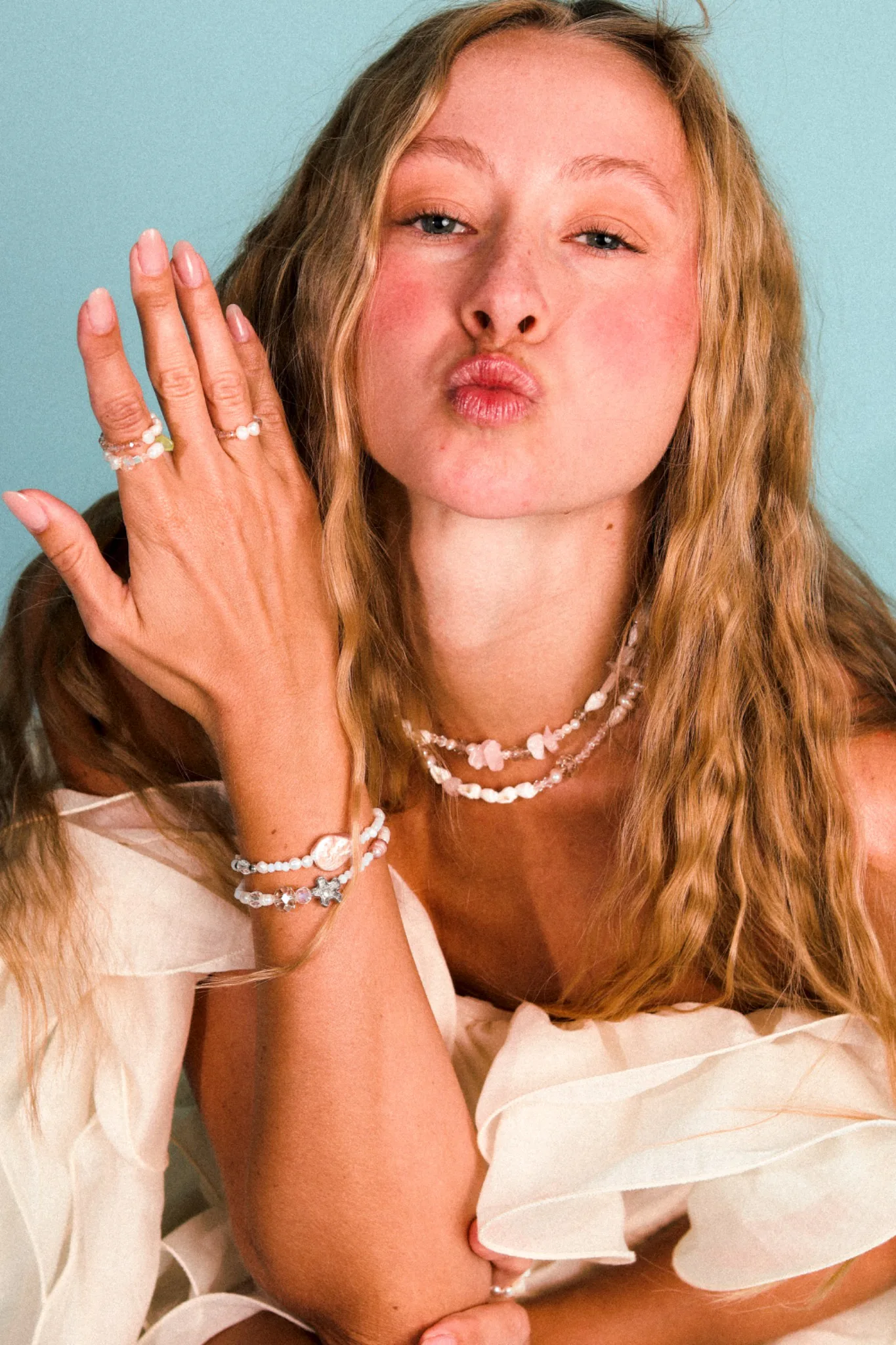 Woman wearing pearl and beaded necklaces, bracelets and rings against a turquoise background