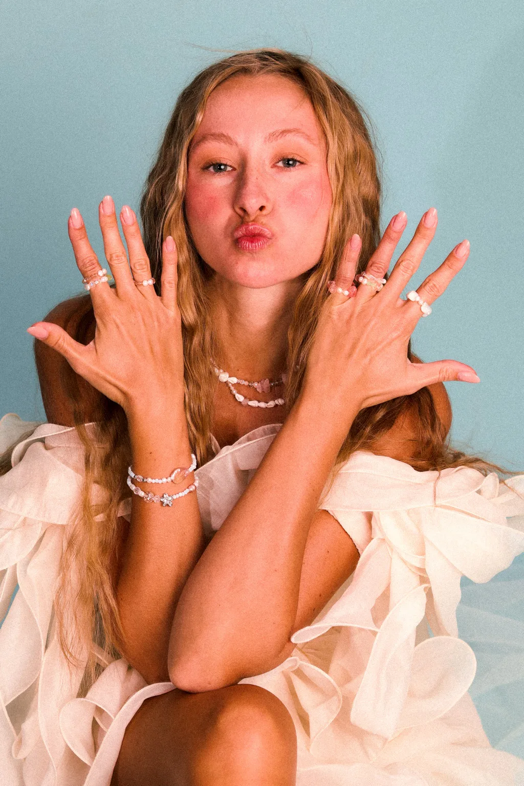 Woman in a white ruffled dress showing pearl rings, bracelets and necklaces against a blue background